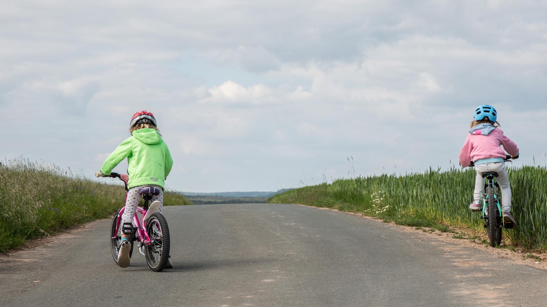 Zwei Kinder mit Helm fahren auf einer leeren Straße mit dem Rad. Eines droht in das Feld abzudriften.