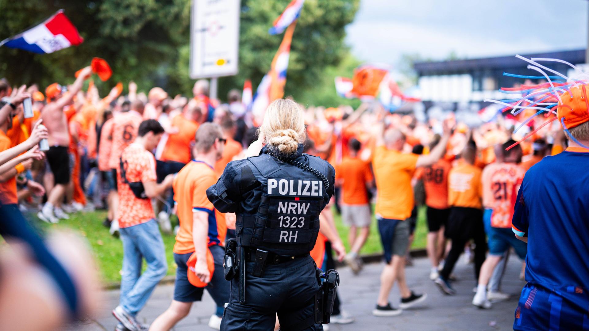 Eine Polizistin steht im Vordergrund; man sieht sie von hinten. Dahinter unscharf eine große Menge an Fans in orangebarbenen Trikots, die vorbeilaufen. 