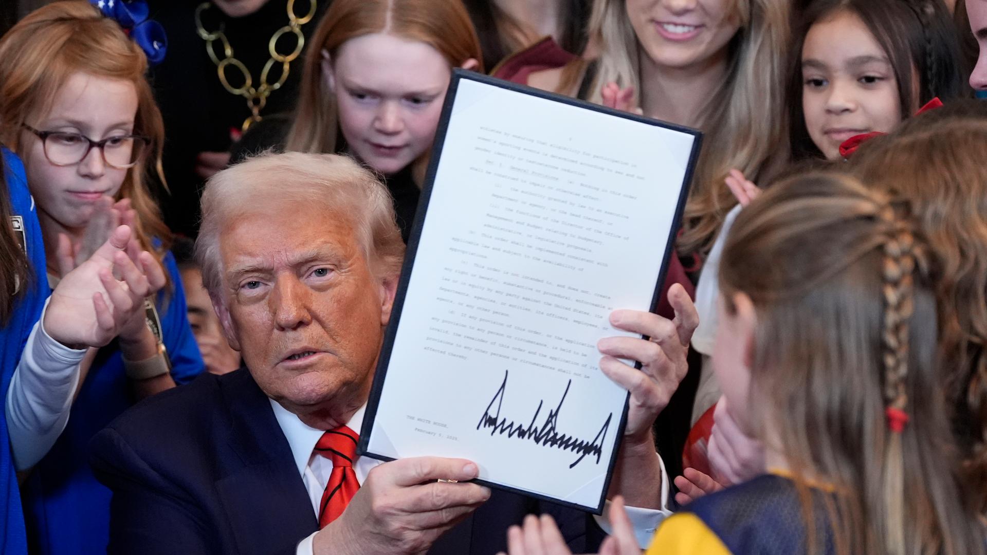 US-Präsident Donald Trump unterzeichnet im Weißen Haus eine Durchführungsverordnung, die transsexuellen Sportlerinnen die Teilnahme an Frauen- und Mädchensportveranstaltungen untersagt. Hinter ihm stehen viele junge Mädchen und Frauen.