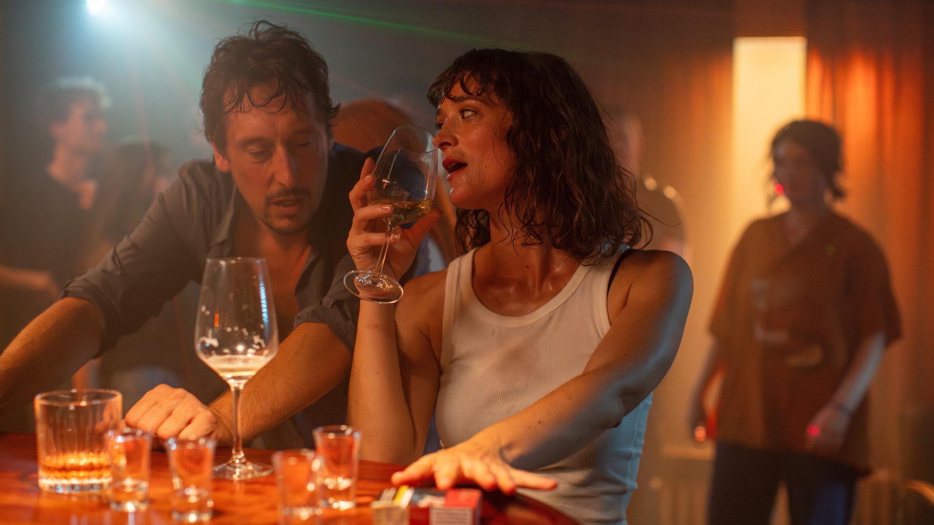 Filmstill aus dem ZDF-Film "Im Rausch". Zu sehen ist eine typische Barszene, der Hintergrund leuchtet rötlich, das Licht ist schummerig. Rechts im Bild eine sichtlich betrunkene Frau, die ein Glas zum Mund führt. Links ein Mann, der auf sie einredet. Filmstill aus dem ZDF-Film "Im Rausch". Zu sehen ist eine typische Barszene, der Hintergrund leuchtet rötlich, das Licht ist schummerig. Rechts im Bild eine sichtlich betrunkene Frau, die ein Glas zum Mund führt. Links ein Mann, der auf sie einredet.