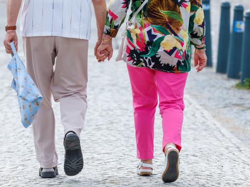 Das Foto zeigt die Beine eines älteren Paares. Beide gehen auf dem Straßenpflaster und tragen Sandalen. Am oberen Bildrand ist zu sehen, dass die beiden Hand in Hand gehen.
