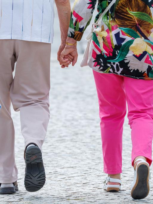 Das Foto zeigt die Beine eines älteren Paares. Beide gehen auf dem Straßenpflaster und tragen Sandalen. Am oberen Bildrand ist zu sehen, dass die beiden Hand in Hand gehen.
