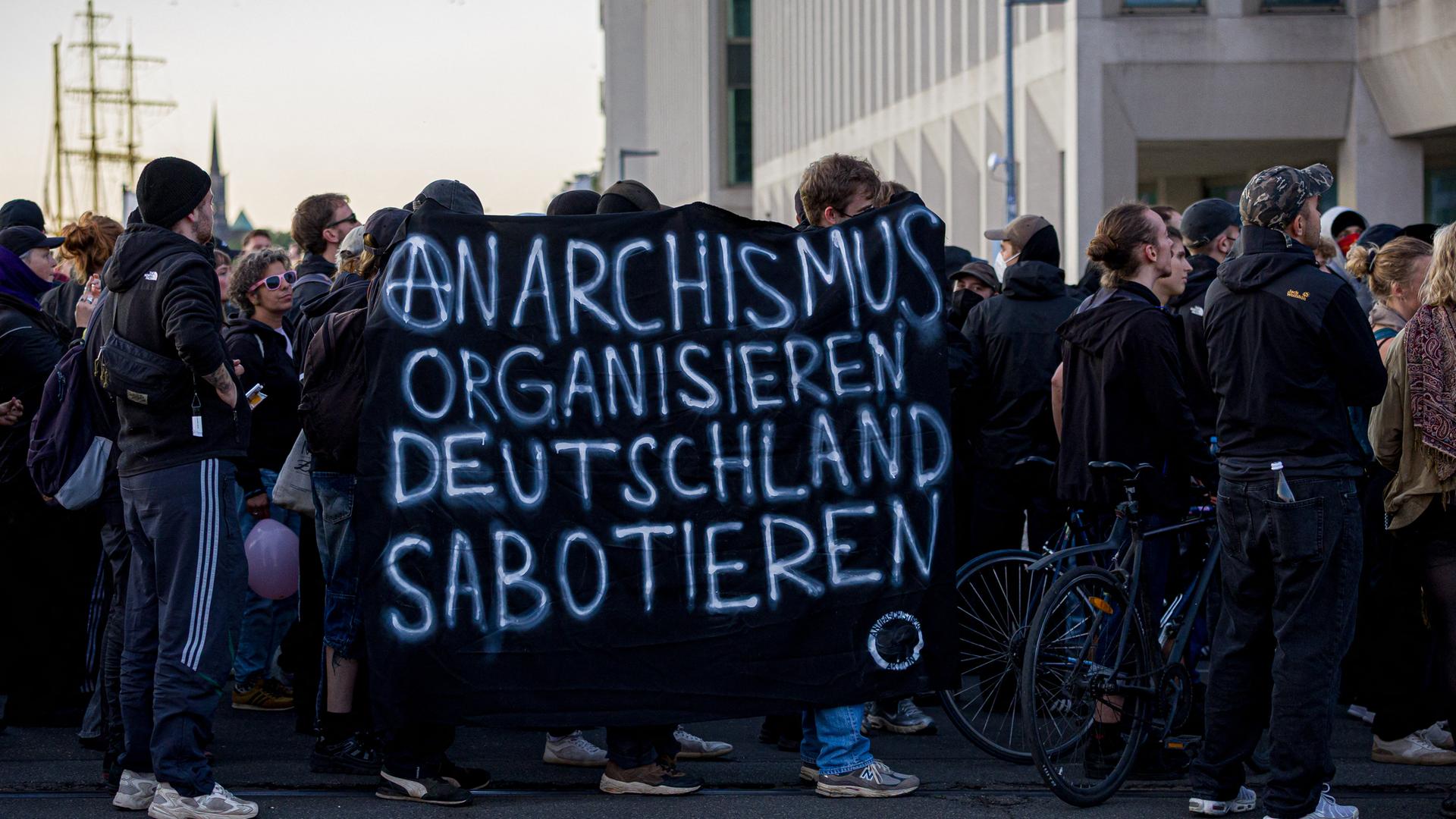 Das Foto zeigt Menschen, die gegen den Tag der Deutschen Einheit demonstrieren. Auf einem Banner ist der Schriftzug "Anarchismus organisieren - Deutschland sabotieren" zu lesen.