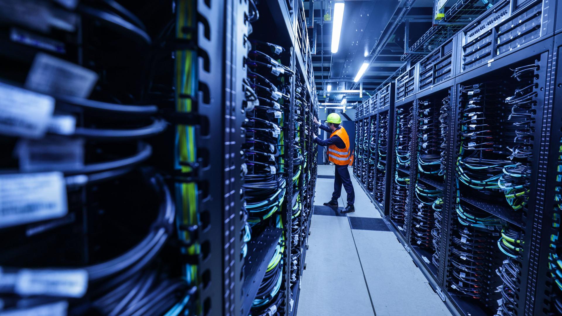 Ein Mitarbeiter mit orange farbener Warnweste iund Sicherheitshelm arbeitet in einem Container des Supercomputers Jupiter im Forschungszentrum Jülich. Ein Mitarbeiter mit orange farbener Warnweste iund Sicherheitshelm arbeitet in einem Container des Supercomputers Jupiter im Forschungszentrum Jülich.