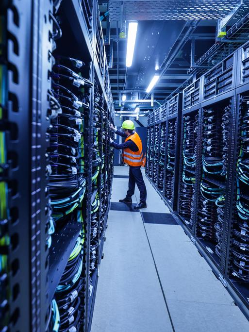 Ein Mitarbeiter mit orange farbener Warnweste iund Sicherheitshelm arbeitet in einem Container des Supercomputers Jupiter im Forschungszentrum Jülich. Ein Mitarbeiter mit orange farbener Warnweste iund Sicherheitshelm arbeitet in einem Container des Supercomputers Jupiter im Forschungszentrum Jülich.