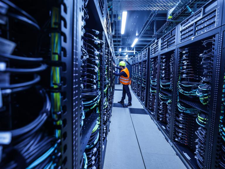 Ein Mitarbeiter mit orange farbener Warnweste iund Sicherheitshelm arbeitet in einem Container des Supercomputers Jupiter im Forschungszentrum Jülich. Ein Mitarbeiter mit orange farbener Warnweste iund Sicherheitshelm arbeitet in einem Container des Supercomputers Jupiter im Forschungszentrum Jülich.