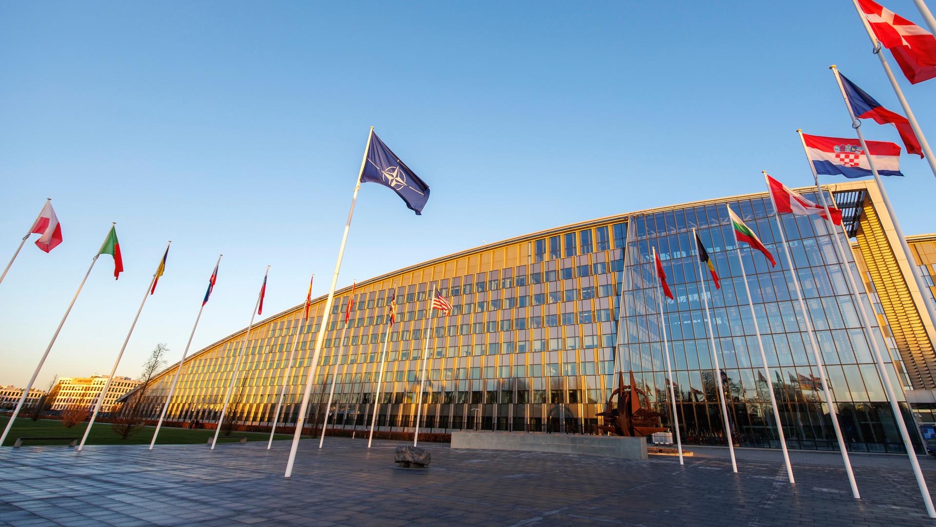 Blick auf das bronzefarbene Gebäude aus Glas und Stahl. Davor Fahnenstangen mit den Flaggen der NATO-Staaten. Der Himmel darüber ist blau. 