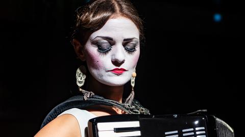 Musikerin Solomiya Melny spielt im Sitzen Akkordeon. Ihr Gesicht ist weiß angemalt, sie trägt ein weißes, ärmelloses Oberteil.