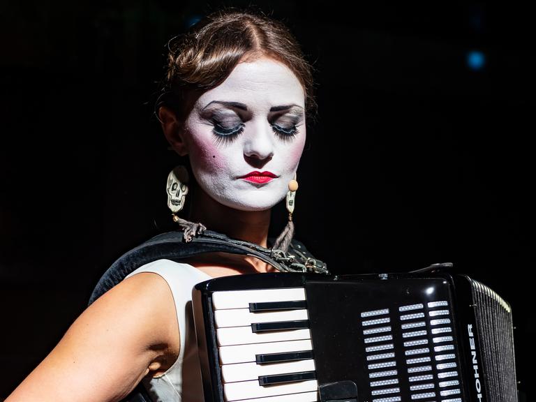 Musikerin Solomiya Melny spielt im Sitzen Akkordeon. Ihr Gesicht ist weiß angemalt, sie trägt ein weißes, ärmelloses Oberteil.