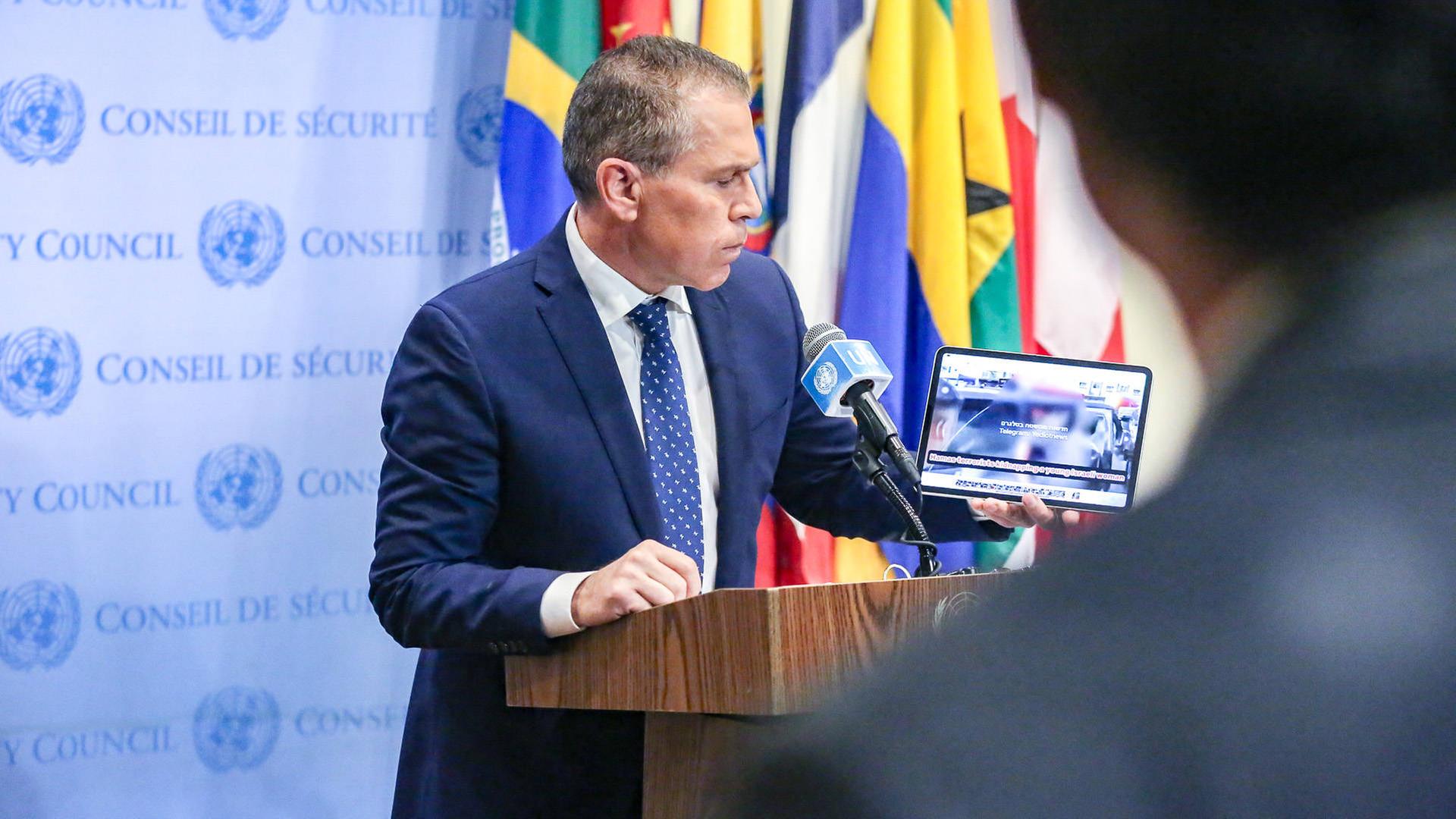 Erdan steht an einem Rednerpult und zeigt etwas auf einem Tablet. Dahinter eine hellblaue Wand mit Logos des UNO-Sicherheitsrates sowie mehrere Flaggen. 