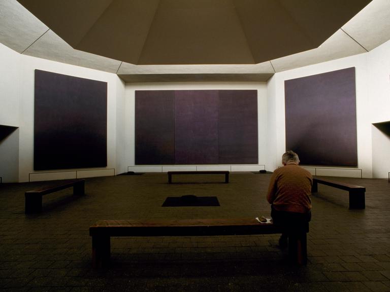 Großflächige schwarze Gemälde an den Wänden eines achteckigen Gebäudes, in der Mitte Holzbänke, darauf ein Mann 