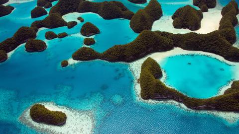Luftaufnahme einer Inselgruppe in Palau: Grün bewaldete Hügel erheben sich aus dem Wasser, in Küstennähe sind Korallenriffe zu sehen.