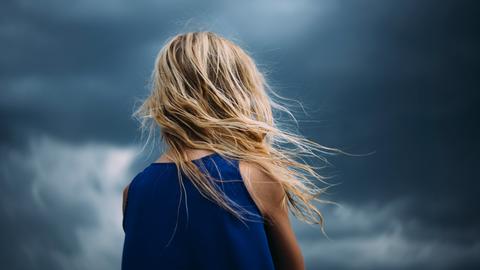 Ein junger Mensch mit langem Haar vor einem Wolkenhimmel.