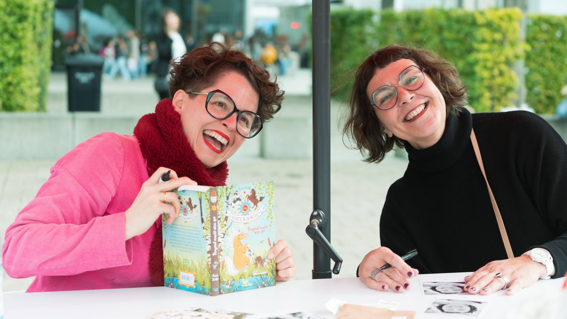 Nina Dulleck und Suza Kolb sitzen gemeinsam an einem Tisch ausserhalb der Messehalle, Nina Dulleck hat das Buch "Haferhorde" in der Hand. Sie lachen ausgelassen.