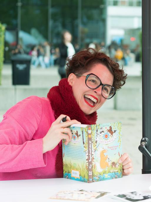 Nina Dulleck und Suza Kolb sitzen gemeinsam an einem Tisch ausserhalb der Messehalle, Nina Dulleck hat das Buch "Haferhorde" in der Hand. Sie lachen ausgelassen.