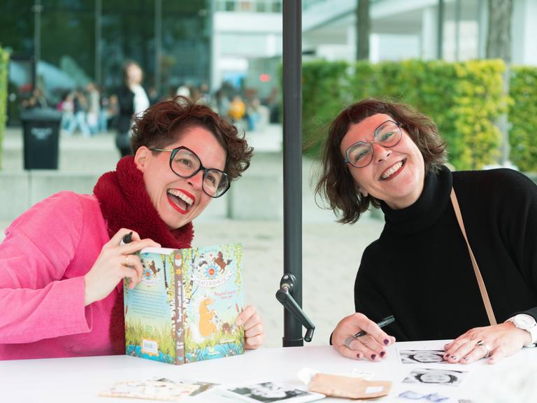 Nina Dulleck und Suza Kolb sitzen gemeinsam an einem Tisch ausserhalb der Messehalle, Nina Dulleck hat das Buch "Haferhorde" in der Hand. Sie lachen ausgelassen.