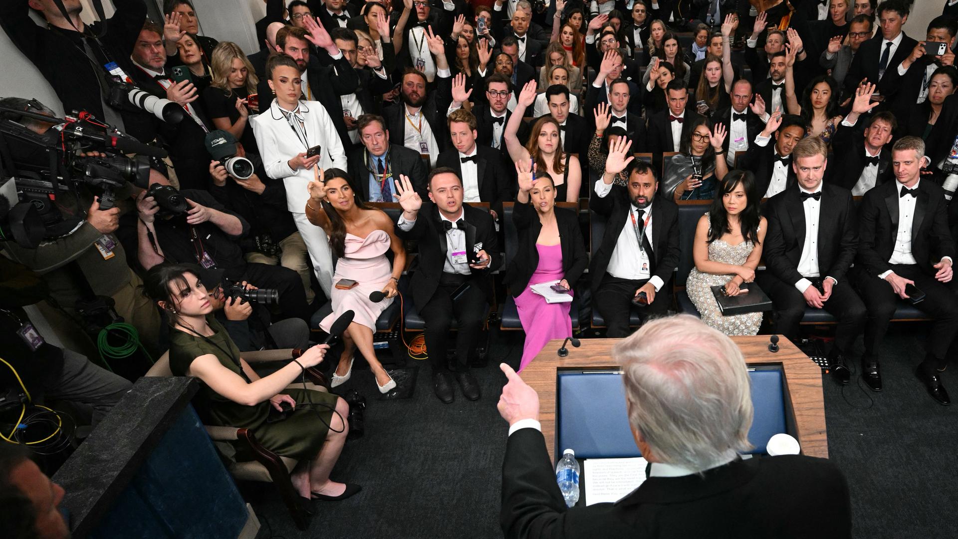 Der Hinterkopf von US-Präsident Donald Trump im Vordergrund, der mit einem Finger auf die vor ihm sitzende Presse zeigt. Viele der Pressevertreter tragen Abendgarderobe und heben die Hand, um eine Frage stellen zu dürfen.