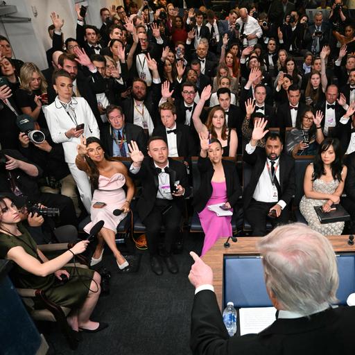 Der Hinterkopf von US-Präsident Donald Trump im Vordergrund, der mit einem Finger auf die vor ihm sitzende Presse zeigt. Viele der Pressevertreter tragen Abendgarderobe und heben die Hand, um eine Frage stellen zu dürfen.