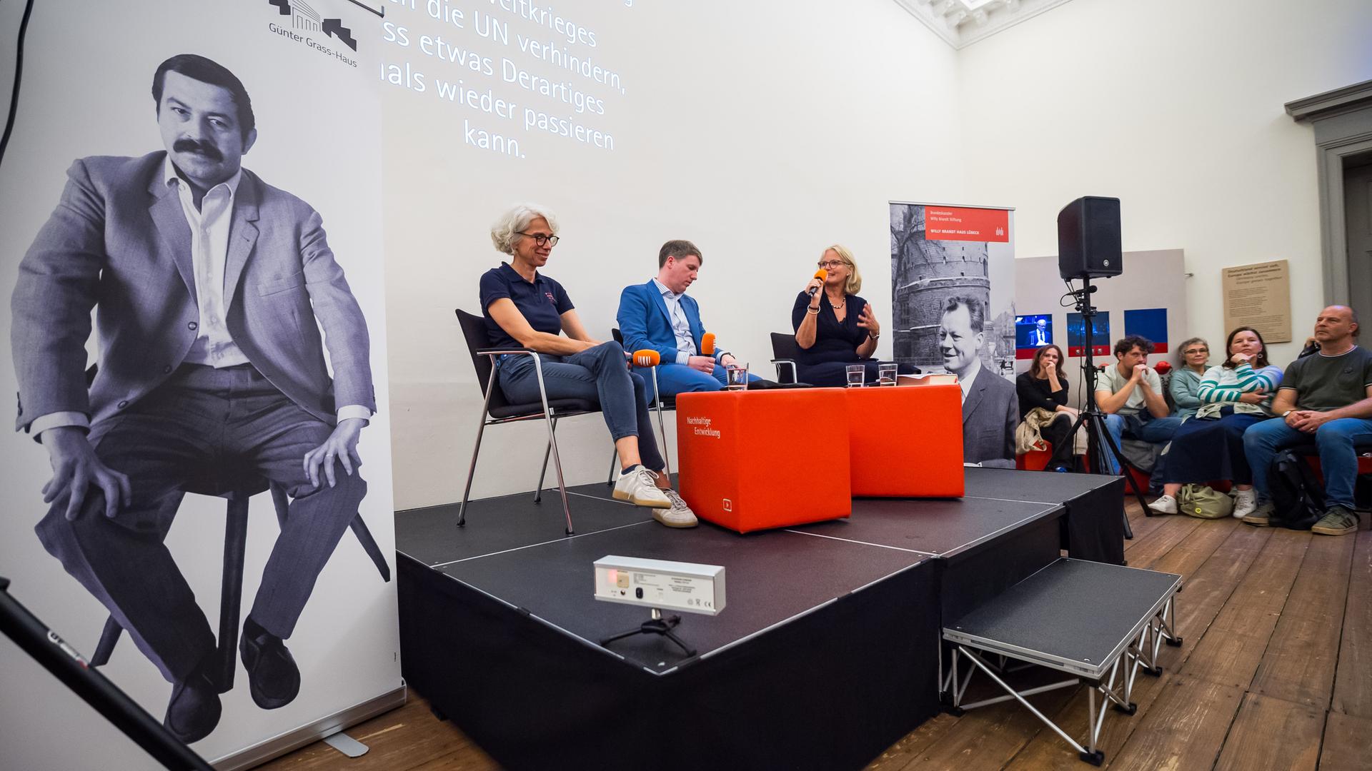 Drei Personen sitzen während einer Veranstaltung auf einem Podium. Sie halten Mikrofone vom Deutschlandradio in den Händen.