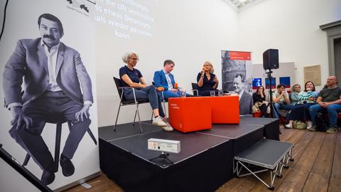 Drei Personen sitzen während einer Veranstaltung auf einem Podium. Sie halten Mikrofone vom Deutschlandradio in den Händen.