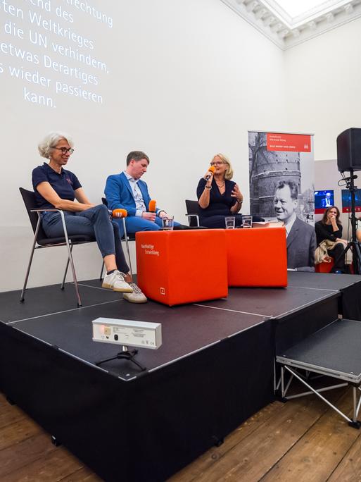 Drei Personen sitzen während einer Veranstaltung auf einem Podium. Sie halten Mikrofone vom Deutschlandradio in den Händen. Drei Personen sitzen während einer Veranstaltung auf einem Podium. Sie halten Mikrofone vom Deutschlandradio in den Händen.