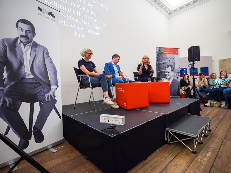 Drei Personen sitzen während einer Veranstaltung auf einem Podium. Sie halten Mikrofone vom Deutschlandradio in den Händen. Drei Personen sitzen während einer Veranstaltung auf einem Podium. Sie halten Mikrofone vom Deutschlandradio in den Händen.