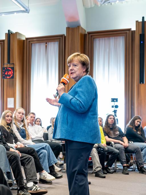 14.10.2026 Aufzeichnung der ersten Folge der Reihe „Merkel unplugged“. Angela Merkel steht vor Schülerinnen und Schülern und hat ein Mikrofon in der Hand. 14.10.2026 Aufzeichnung der ersten Folge der Reihe „Merkel unplugged“. Angela Merkel steht vor Schülerinnen und Schülern und hat ein Mikrofon in der Hand.