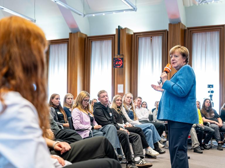 14.10.2026 Aufzeichnung der ersten Folge der Reihe „Merkel unplugged“. Angela Merkel steht vor Schülerinnen und Schülern und hat ein Mikrofon in der Hand. 14.10.2026 Aufzeichnung der ersten Folge der Reihe „Merkel unplugged“. Angela Merkel steht vor Schülerinnen und Schülern und hat ein Mikrofon in der Hand.