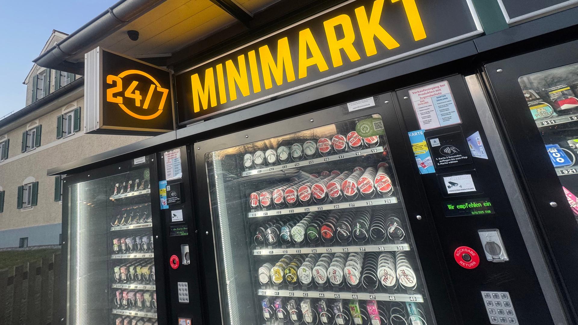 Blick schräg von der Seite auf die Front eines "Minimarkt", in dem man Produkte in einem Verkaufsautomat sieht. 