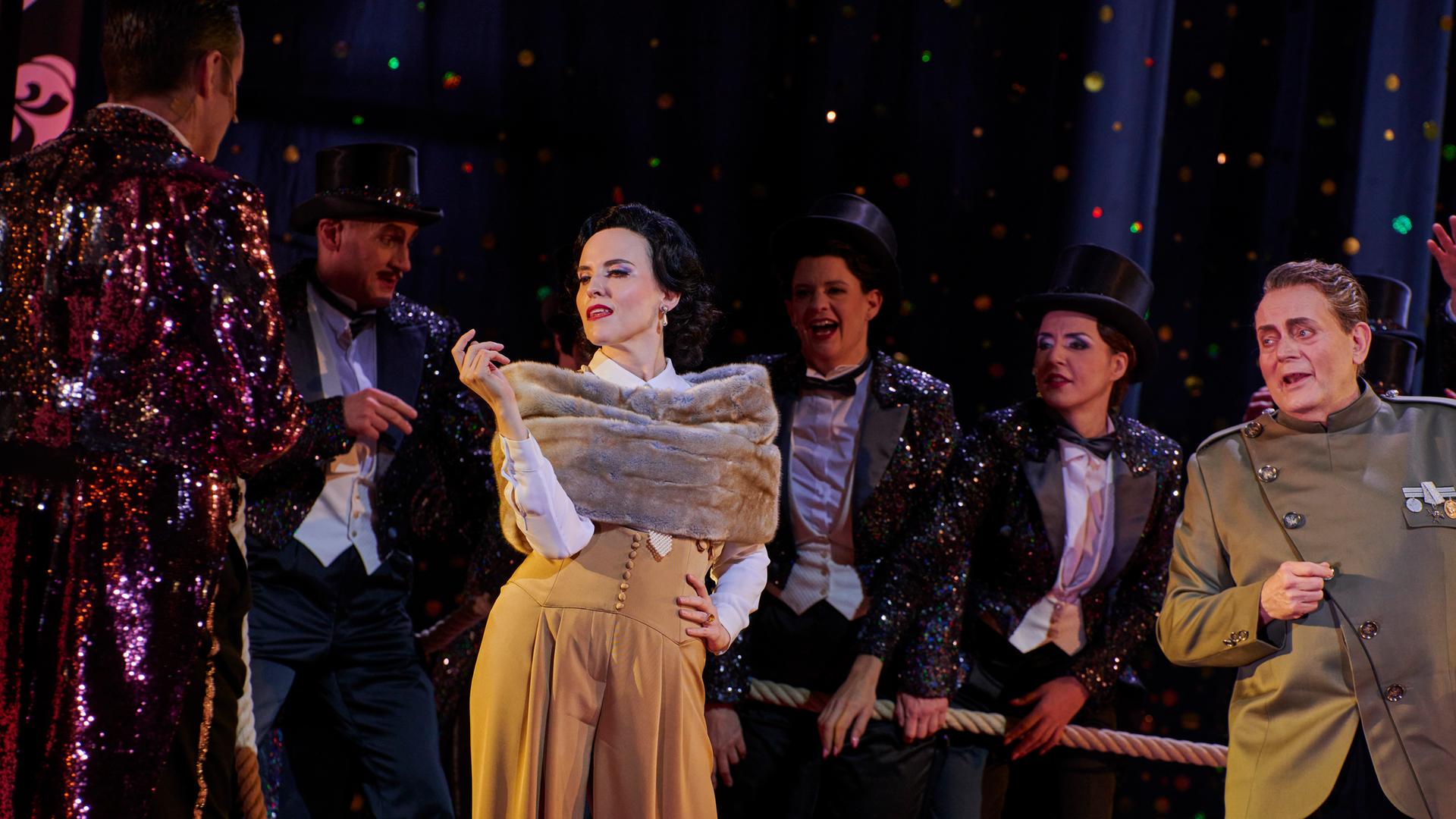 Szenenbild der Jazz-Operette "Die lustige Witwe": Die Schauspielerin Rebecca Nelsen posiert in ihrer Rolle als Hanna Glawari in luxuriös anmutender Kleidung vor dem Ensemble. 