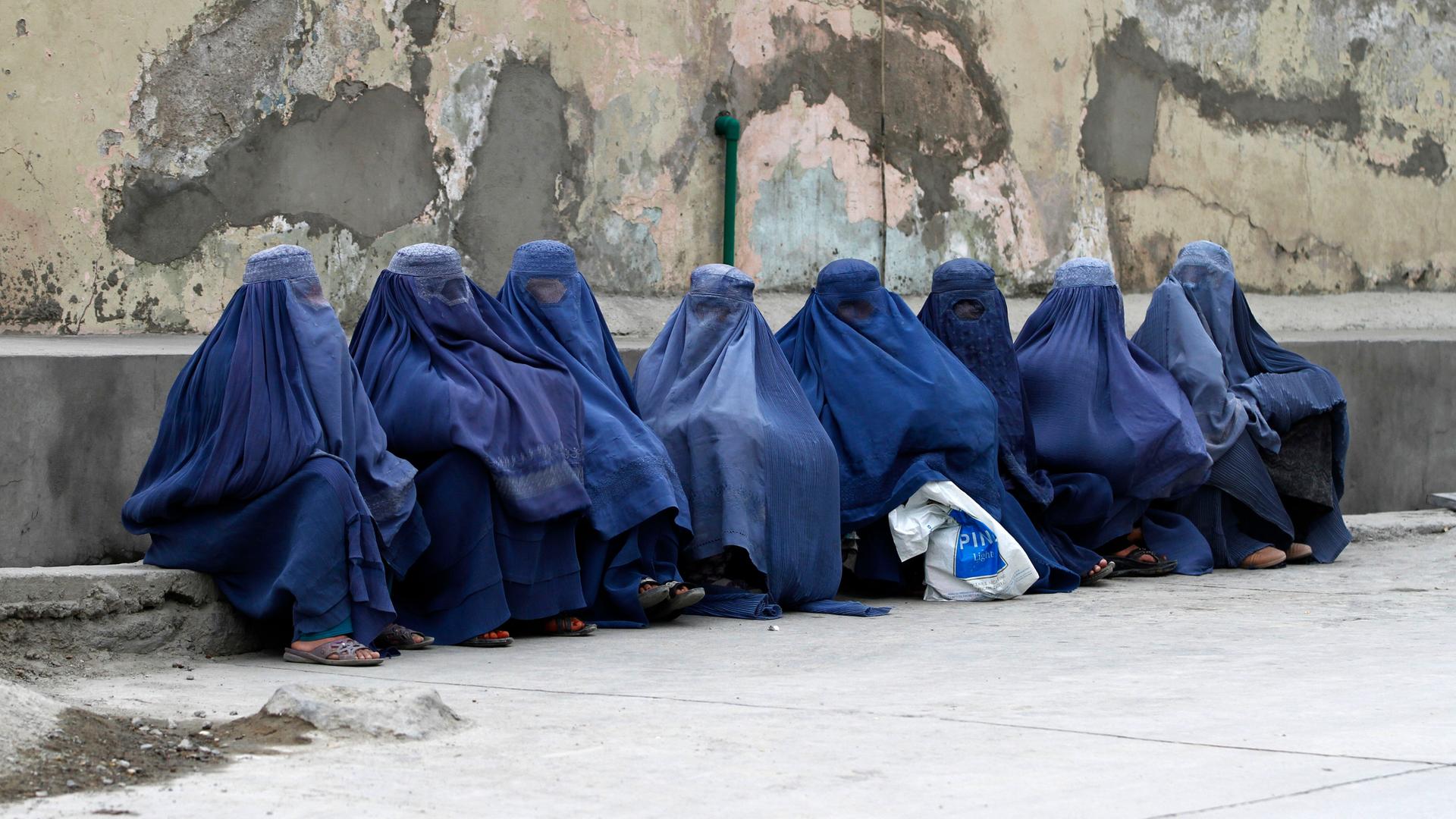 Mehrere afghanischen Frauen sitzen komplett verschleiert durch Burkas auf Rinnstein an der Straße. 