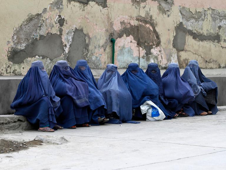 Mehrere afghanischen Frauen sitzen komplett verschleiert durch Burkas auf Rinnstein an der Straße. 
