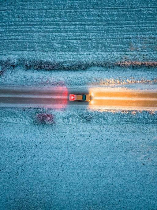 Vogelperspektive auf ein Auto, das im Dunkeln durch eine Schneelandschaft fährt.
