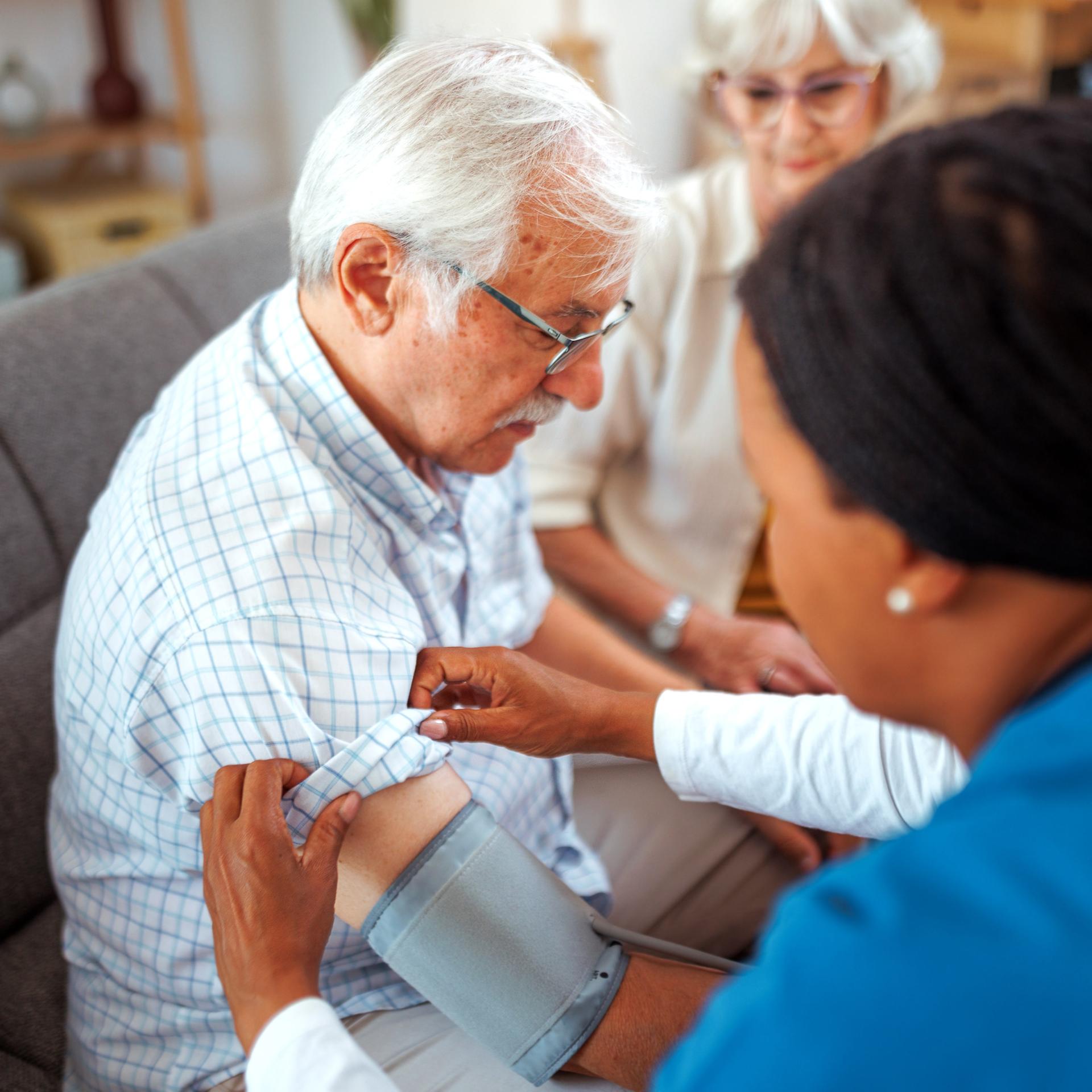 Eine Krankenschwester checkt bei einem Hausbesuch den Blutdruck eines älteren Herren.