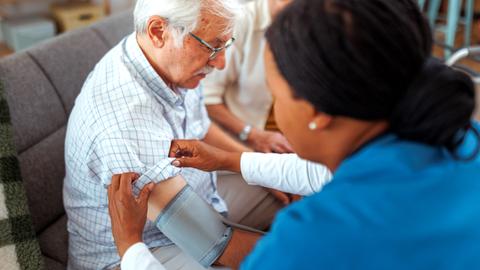 Eine Krankenschwester checkt bei einem Hausbesuch den Blutdruck eines älteren Herren.