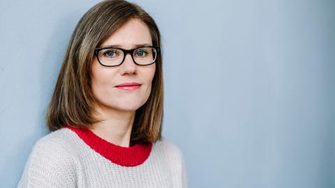 Eine Frau mit braunen schulterlangen Haaren, einer Brille und einem Strickpullover mit roten Rand blickt vor einem blau-grauen Hintergrund in die Kamera.