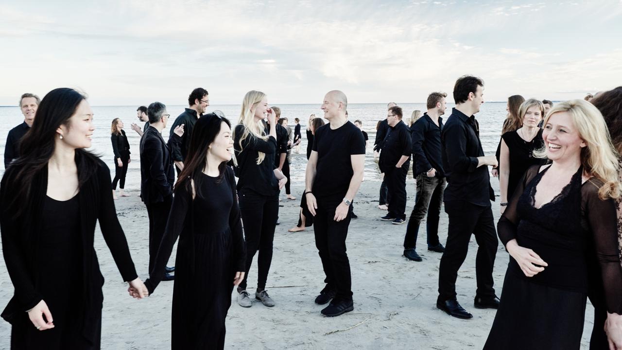 Eine große Gruppe schwarz gekleideter Frauen und Männer stehen in vergnügter Laune verstreut an einem Strand zusammen.
