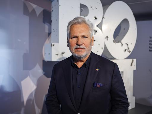 Polens früherer Staatschef Aleksander Kwaśniewski steht in einem dunklen Anzug vor einer Wand auf der große Buchstaben zu sehen sind