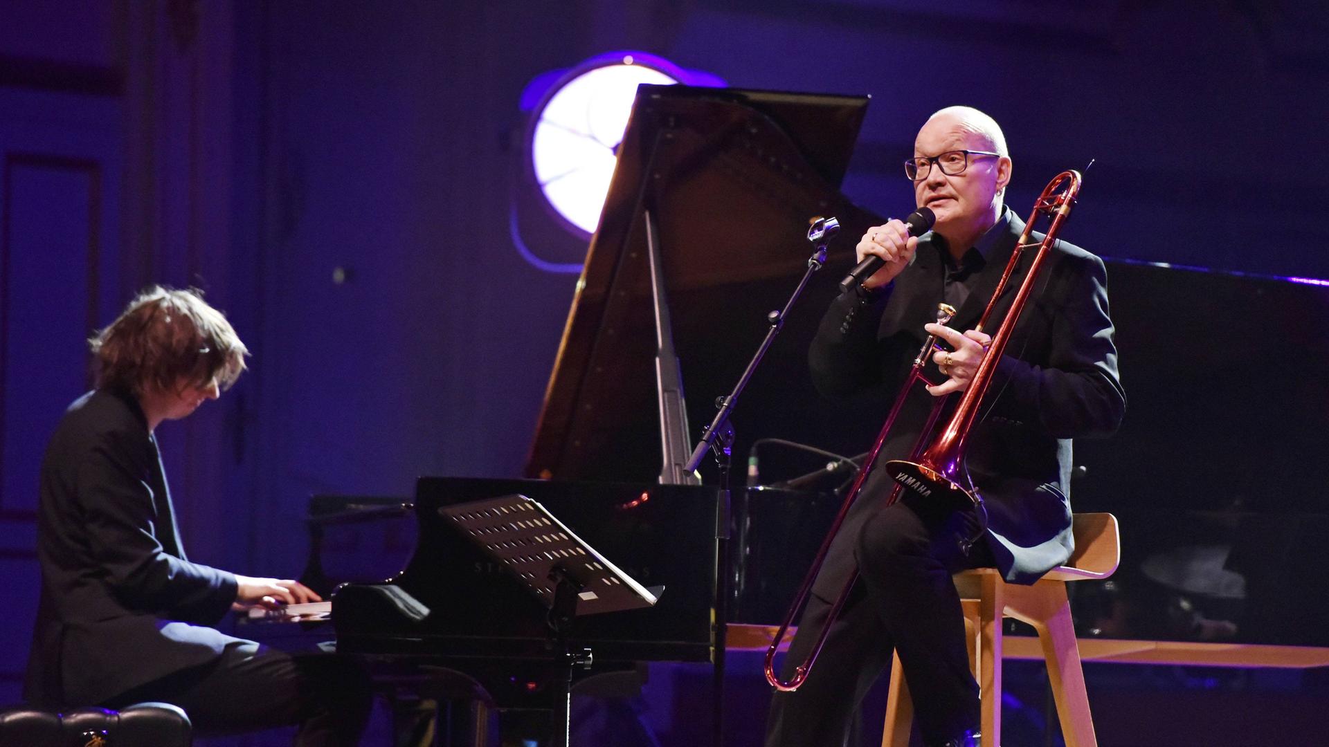 Michael Wollny begleitet Nils Landgren (Posaune) am Klavier auf einer rot-blau ausgeleuchteten Bühne in Hamburg im Jahr 2019. 