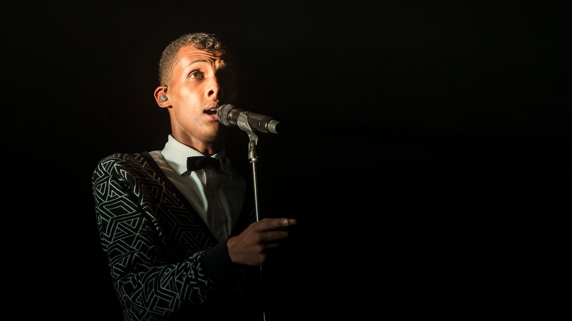Der Sänger Stromae bei einem Konzert 2014: Er steht vor einem Mikro, der Hintergrund ist schwarz.