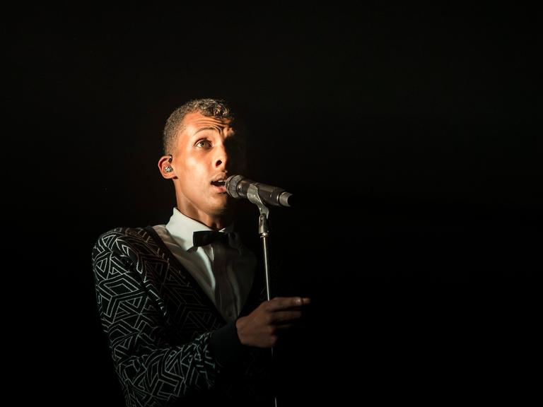 Der Sänger Stromae bei einem Konzert 2014: Er steht vor einem Mikro, der Hintergrund ist schwarz.