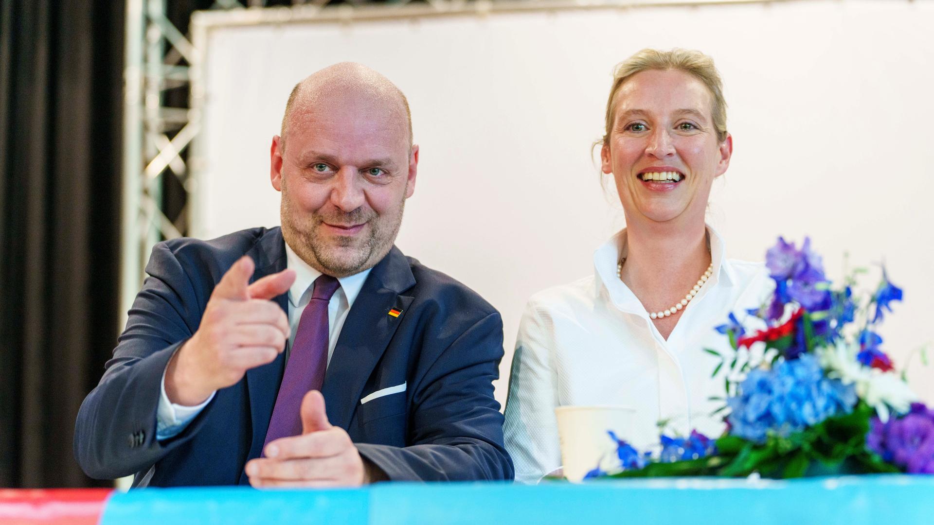 Hessens AfD-Spitzenkandidat Robert Lambrou zeigt lächelnd Richtung Kamera, neben ihm sitzt lachend die Parteivorsitzende Alice Weidel