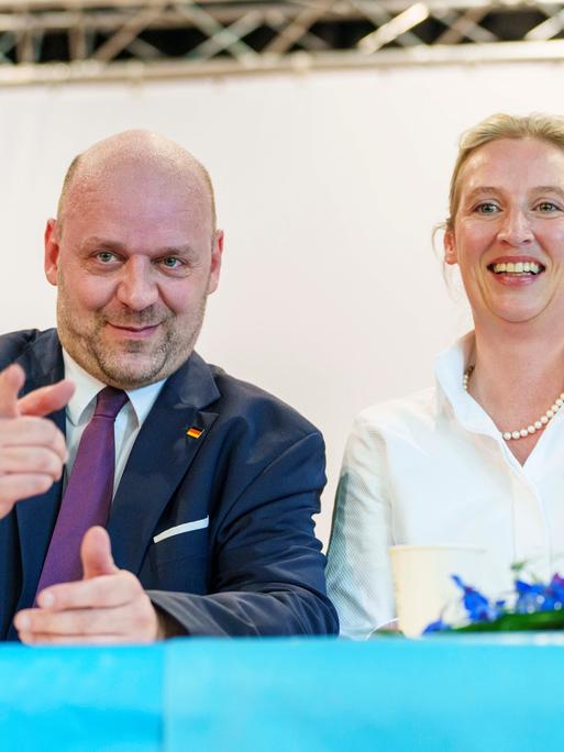 Hessens AfD-Spitzenkandidat Robert Lambrou zeigt lächelnd Richtung Kamera, neben ihm sitzt lachend die Parteivorsitzende Alice Weidel