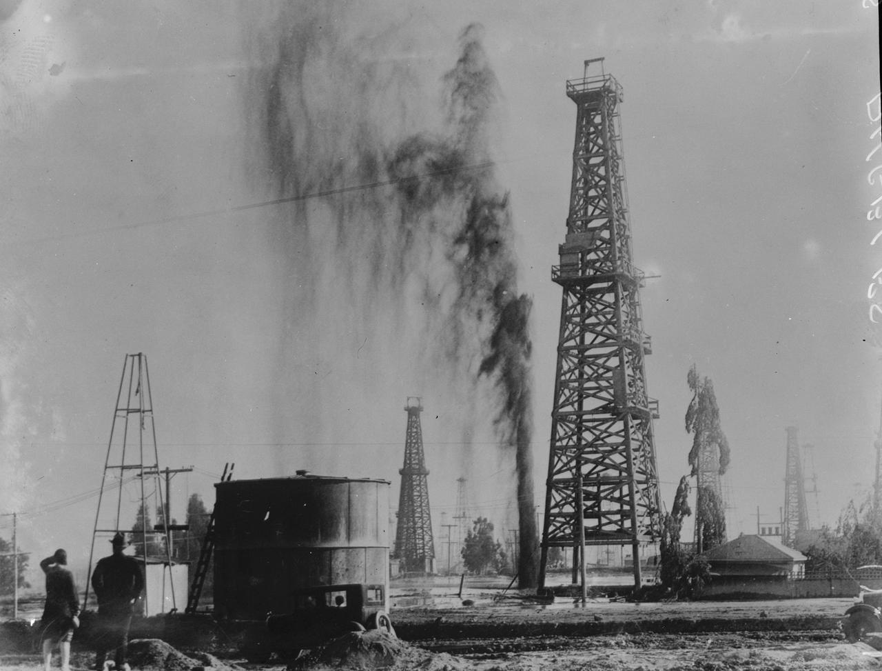 Auf dem Schwarz-Weiß-Foto ist ein Ölfeld zu sehen, auf dem eine Ölfontäne in den Himmel schießt.