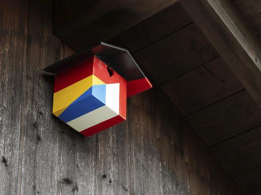 Ein Nistkasten für Vögel in den Farben der deutschen und französischen Nationalflagge hängt an unter einem hölzernen Dachgiebel. 
