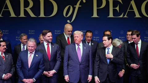 Die drei und weitere Teilnehmer stehen neben- und hintereinander auf einer Bühne. Dahinter eine blaue Wnnd mit dem goldenen Schriftzug "Board of Peace".