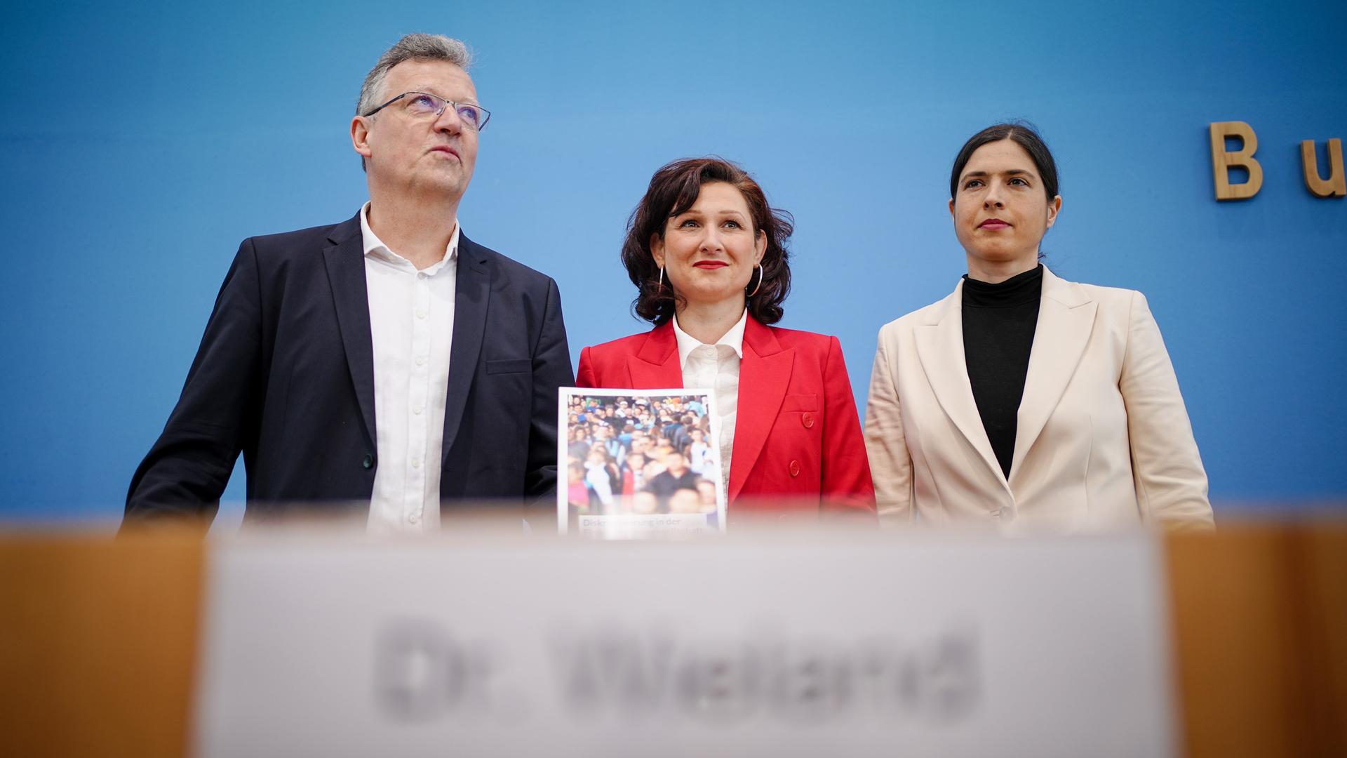 Die drei stehen nebeneinander hinter einem Pult vor der blauen Wand der Bundespressekonferenz. Ataman in der Mitte im roten Jackett hält die Studie. Die drei stehen nebeneinander hinter einem Pult vor der blauen Wand der Bundespressekonferenz. Ataman in der Mitte im roten Jackett hält die Studie.