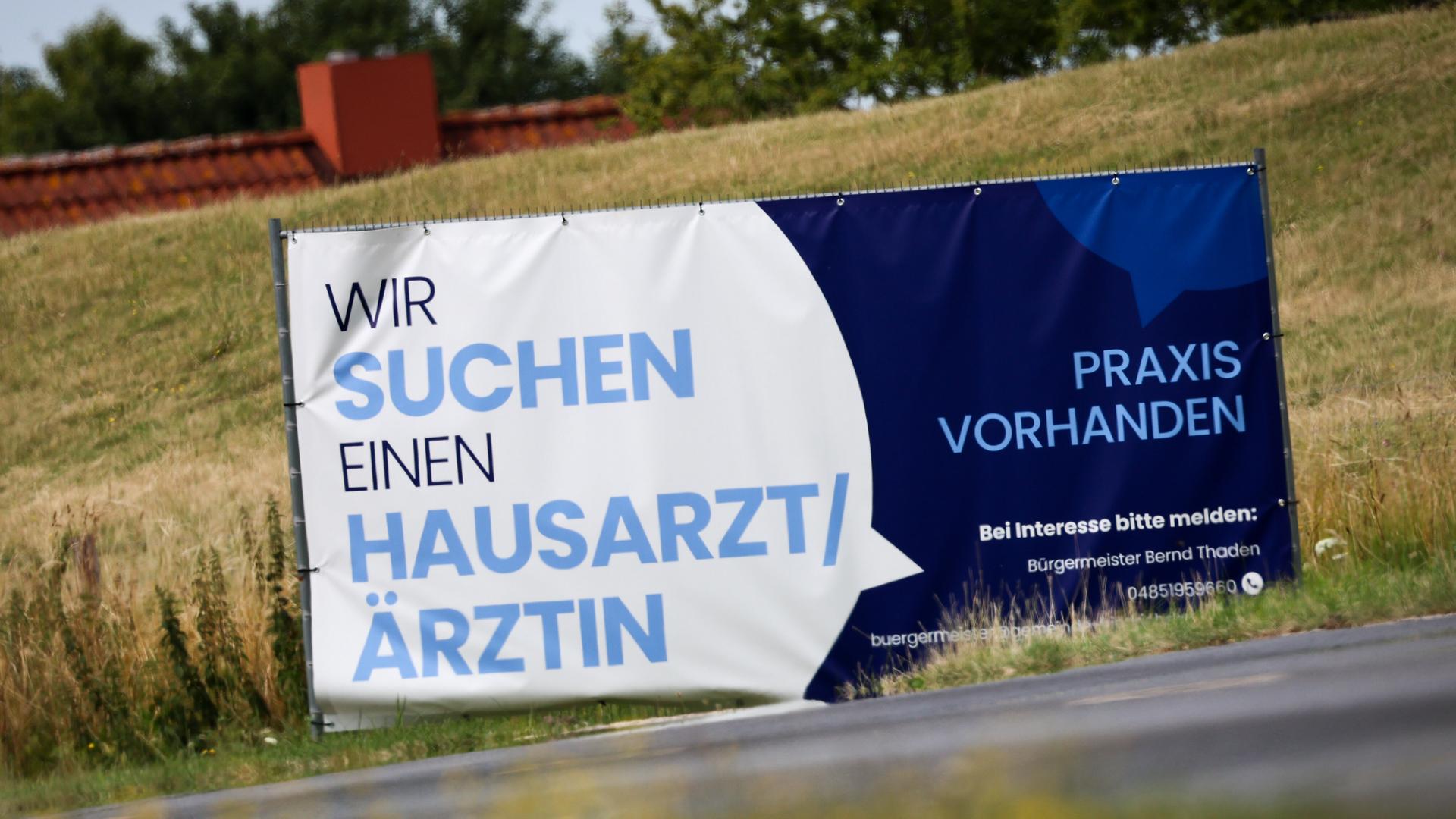 Ein Plakat mit dem Text "Wir suchen einen Hausarzt / Ärztin - Praxis Vorhanden" steht an einer Straße 