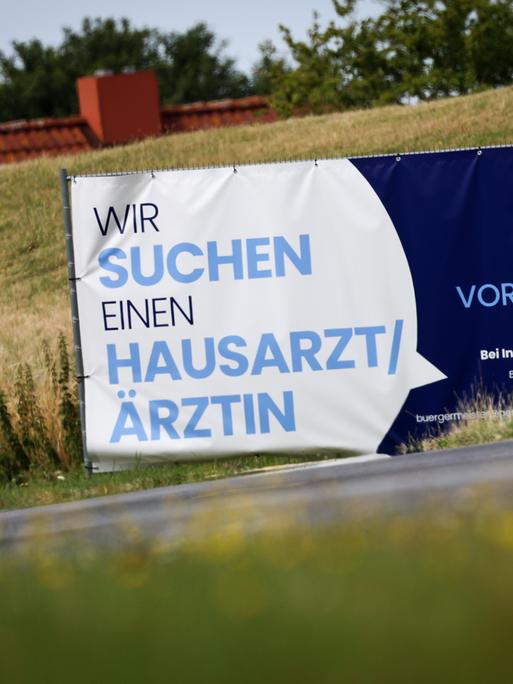 Ein Plakat mit dem Text "Wir suchen einen Hausarzt / Ärztin - Praxis Vorhanden" steht an einer Straße 