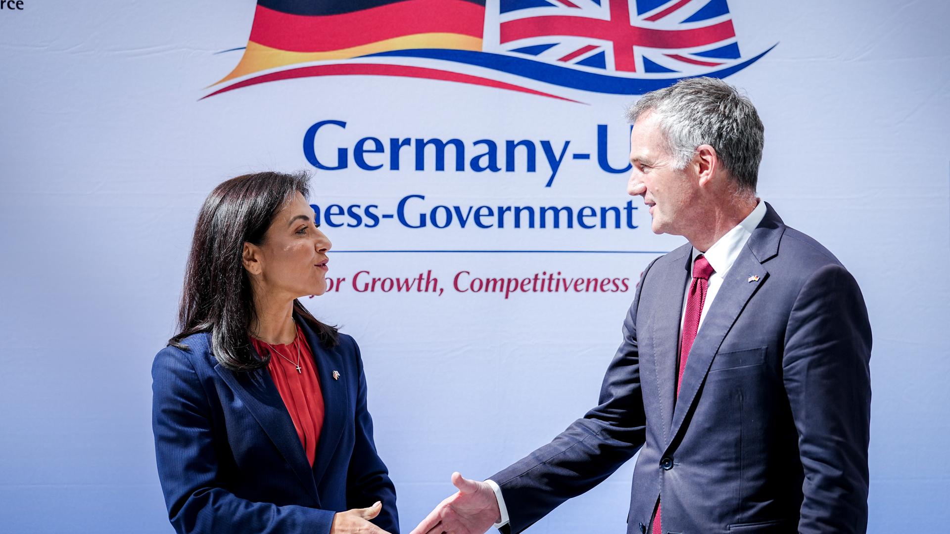 Reiche begrüßt Kyle mit Handschlag. Beide schauen sich an. Im Hintergrund sind die Flaggen beider Länder zu sehen. 